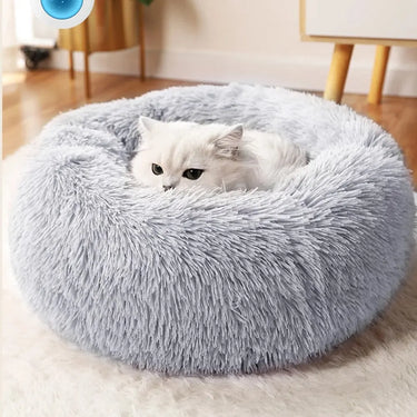 A fluffy white cat lounges in the KittyNook Cat Co. Warm & Soft Cat Sleeping Pod, a round plush light gray bed.